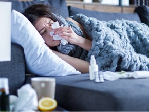 Sick exhausted girl in scarf is lying in bed sofa wrapped in grey blanket. Young woman with runny nose, headache and fever is blowing out snot. Home treatment. Winter cold and flu concept.